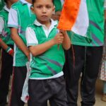 Young boy in a green school uniform holds an orange and white flag at a line of teammates behind him in a school event